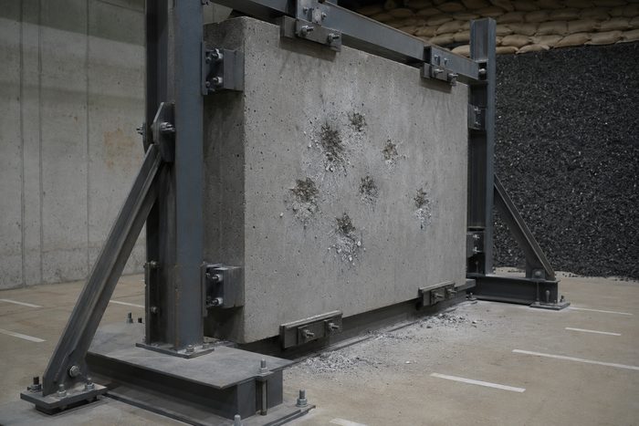 Concrete test panel clamped in a steel test frame at an indoor range, its face marked with ballistic impact craters